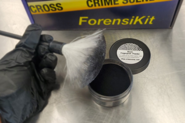 Gloved hand holding a fingerprint brush over a vial of fingerprint powder with the ForensiKit box in the background